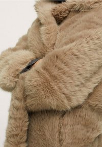 Cappotto in pelliccia sintetica di colore marrone chiaro con una texture morbida e pelosa. Presenta un colletto rotondo e dettagli ai polsi, evidenziando un design accogliente e spesso.