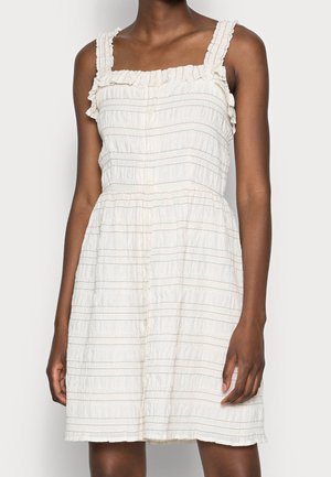 Kobieta w białej sukience bez rękawów z marszczonymi ramiączkami i poziomymi teksturowanymi paskami, stojąca na jednolitym jasnym tle.