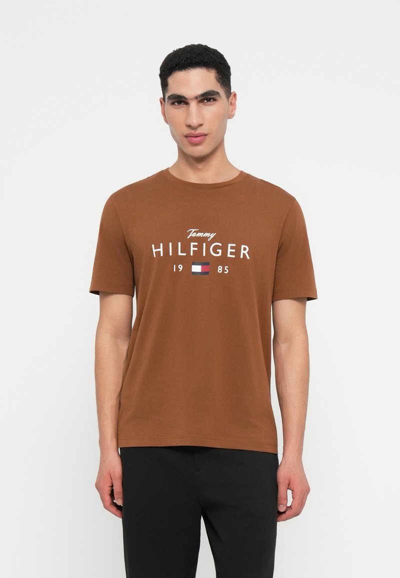 Muž v hnědém tričku Tommy Hilfiger s logem a černých kalhotách, stojící na pozadí s jednoduchou bílou texturou.