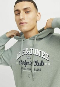Felpa grigia in cotone con cappuccio regolabile, caratterizzata da testo stampato in blu navy e bianco, inclusa la dicitura "JACK&JONES Surfer's Club." Texture morbida.