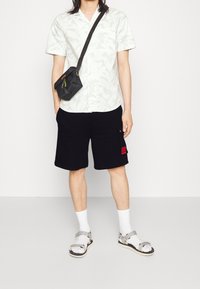 Camisa de manga corta blanca con estampado floral; pantalones cortos cargo negros; bolso cruzado negro; calcetines blancos con sandalias; corte holgado.