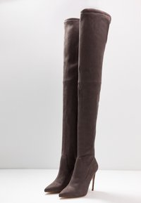 Stivali alti in suede marrone scuro con punte appuntite e tacchi a stiletto, poggianti in posizione verticale su una superficie bianca.