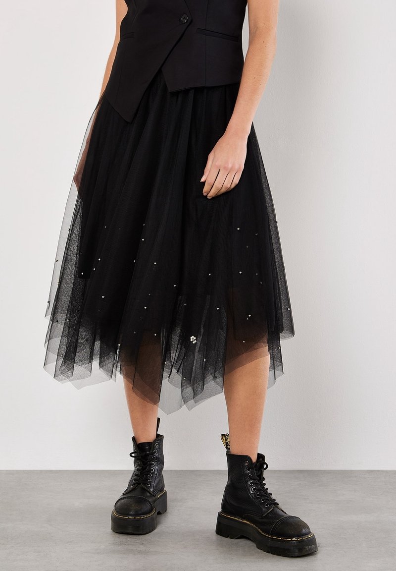 Femme portant une jupe en tulle noire superposée avec de petites perles blanches et des bottines noires à lacets sur plateforme, debout sur un sol gris.