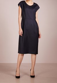 Námořnické modré šaty s krátkým rukávem a kulatým výstřihem, po kolena, s bočními kapsami a hladkým texturovaným materiálem. Doplněné o černé podpatky.