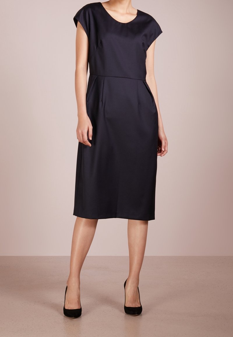 Námořnické modré šaty s krátkým rukávem a kulatým výstřihem, po kolena, s bočními kapsami a hladkým texturovaným materiálem. Doplněné o černé podpatky.