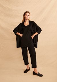 Zwarte loszittende cardigan met wijde mouwen, gecombineerd met een zwarte tanktop en cropped broek. Model draagt zwarte instappers. Getextureerde achtergrond.