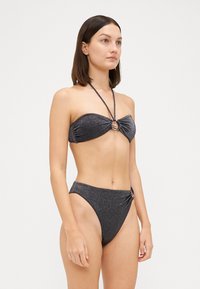 Juodas blizgantis bikinis su bandeau stiliumi, turinčiu centrinį žiedą. Aukšto liemens kelnaitės su šoniniu akcentu. Lygi tekstūra ir elastingas kirpimas.