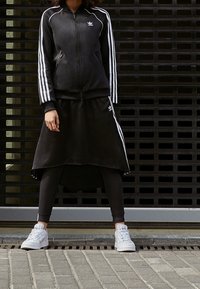 Svart zip-up jacka med vita ränder, kombinerad med en svart kjol och leggings. Vita sneakers fulländar den sportiga looken.