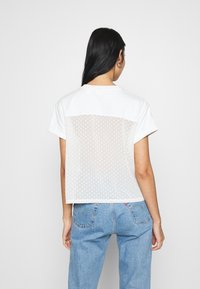 Weißes T-Shirt mit kurzen Ärmeln, durchscheinendem gepunktetem Rückenteil, rundem Ausschnitt und geradem Saum, kombiniert mit blauen Jeans.