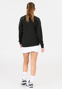 Kvinna med långt hår i hästsvans, iklädd en svart långärmad tröja, vit kjol, vita strumpor och svart-vita sneakers, stående med ryggen vänd.