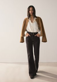 Hnedá semišová bunda cez bielu textúrovanú košeľu s gombíkmi, v kombinácii s čiernymi nohavicami s vysokým pásom a rozšírenými nohavicami a tmavými topánkami. Jednoduchý, moderný dizajn.