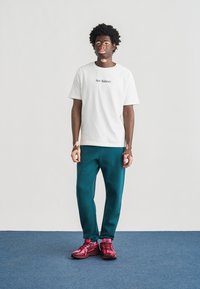 Λευκό βαμβακερό T-shirt με λογότυπο "New Balance", πετρόλ παντελόνια φόρμας και ροζ αθλητικά παπούτσια. Στέκεται σε μπλε χαλί.