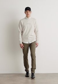 Krémová mikina s rebrovanými manžetami, svetlý košeľový tričko pod ňou, olivovozelené slim-fit nohavice a čierne topánky. Na hlave má štruktúrovanú čiernu čiapku.
