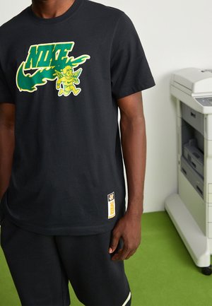 Zwart katoenen T-shirt met een groen en geel Nike-logo en grafische accenten. Heeft een gele tag aan de zijkant. Draagt zwart korte broek.