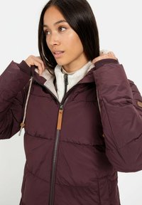 Bordeauxkleurige pufferjack met een rits en een getoggelde capuchon, voorzien van een fleece-gevoerde kraag en borst, en tan leren accenten op de mouwen.