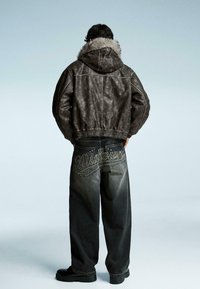 Brun konstläderjacka med en luvtröja fodrad med päls, kombinerad med löst sittande grå denimjeans med "Midtown" broderi på baksidan.
