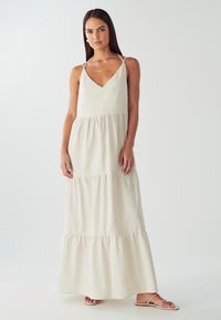 Moteris vilkinti be Rankovių, smėlio spalvos, sluoksniuotą maksi suknelę su špagetėmis dirželis ir plokščiomis sandalais prieš paprastą baltą foną.