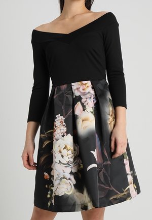 Femme portant un haut noir à épaules dénudées avec des manches trois-quarts et une jupe longueur genou ornée d'un grand imprimé floral.