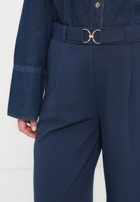 Μαύροι σκούροι μπλε παντελόνες ψηλής μέσης με λεία υφή, συνδυασμένοι με τοπ από τζιν. Τα παντελόνια διαθέτουν λεπτομέρεια με αγκράφα σε χρυσό τόνο στη ζώνη.