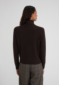 Un maglione a collo alto marrone presenta un colletto e un orlo a costine, realizzato in materiale morbido, abbinato a pantaloni a quadri a vita alta.
