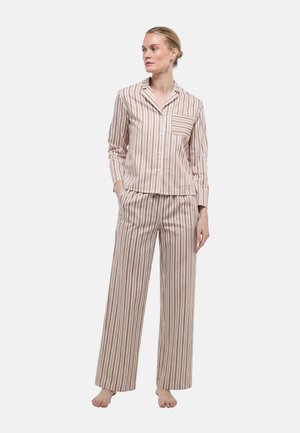 Femme debout, pieds nus, portant une chemise à manches longues à boutons avec des rayures verticales beige et marron, assortie à un pantalon large.