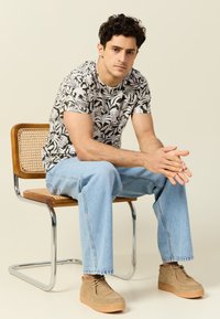 Camisa de manga corta con un patrón de hojas en blanco y negro, jeans de mezclilla azul claro y zapatos de ante beige, sentado en una silla de madera y metal.
