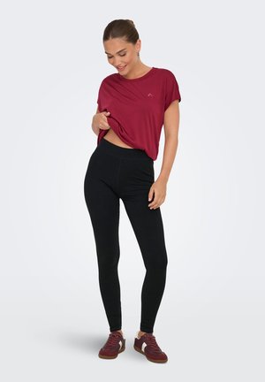 Femme portant un t-shirt bordeaux, des leggings noirs et des baskets bordeaux, regardant vers le bas et soulevant légèrement son t-shirt sur un fond uni.