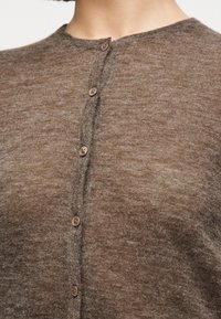 Κοντινό πλάνο ενός ατόμου που φοράει καφέ ζακέτα με κουμπιά και στρογγυλή λαιμόκοψη, εμφανίζοντας το πάνω μέρος του στήθους και την περιοχή του λαιμού.