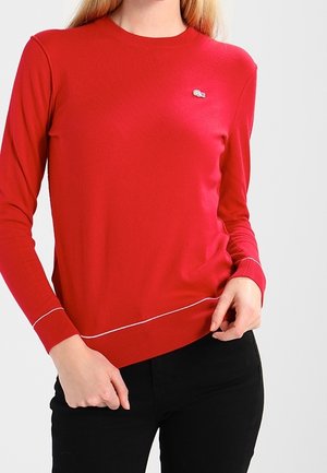 Femme portant un pull rouge à manches longues avec un petit logo sur la poitrine et un pantalon noir, les mains ajustant l'ourlet du pull.