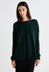 Pulover verde închis din material moale, cu o croială lejeră și un decolteu larg. Mâneci lungi cu manșete și tiv cu nervuri.