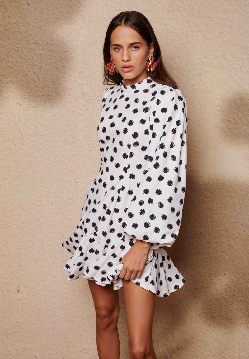 Λευκό φόρεμα με μαύρα πουά, μακριά φουσκωτά μανίκια και βολάν στο κάτω μέρος. Διακριτικά στοιχεία είναι τα τολμηρά κόκκινα σκουλαρίκια με λουλούδια.