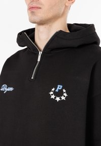 Felpa nera con cappuccio e zip a un quarto, caratterizzata da loghi ricamati blu e bianchi e un motivo a stelle attorno all'area del petto.