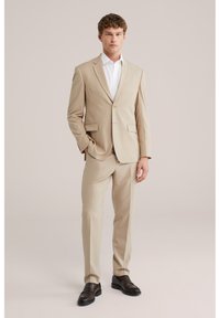 Uomo in piedi con le mani in tasca che indossa un abito beige, camicia bianca e scarpe eleganti marrone scuro su uno sfondo semplice.