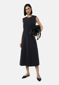 Robe sans manches de couleur marine avec un corsage ajusté et une jupe évasée, présentant une ouverture en forme de clé à l'avant. Associée à un sac noir et des ballerines pointues.