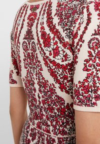 Kleid mit kurzen Ärmeln in Creme, verziert mit filigranen roten Blumen- und Paisleymustern. Hat einen runden Ausschnitt und dezente Nähte in der Taille.