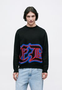 Maglione nero a maglia con bold lettering grafico rosso e blu sulla parte anteriore. Il design ha un classico scollo a girocollo e polsini a coste.