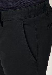 Nahaufnahme einer schwarzen Hosentasche und des Hosenbundes mit sichtbaren Nähten an einer Person, die einen dunkelblauen Strickpullover trägt.