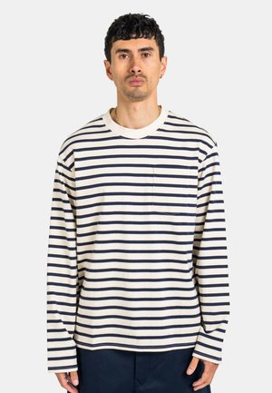 Man wearing a white long-sleeve shirt with black horizontal stripes and a chest pocket, standing against a plain light background.
