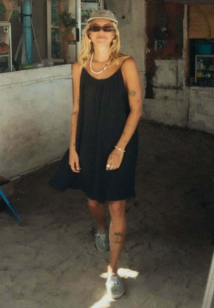 Kobieta w czarnej sukience bez rękawów, zielonych trampkach, beżowej czapce i okularach przeciwsłonecznych stojąca wewnątrz na piaszczystej podłodze.