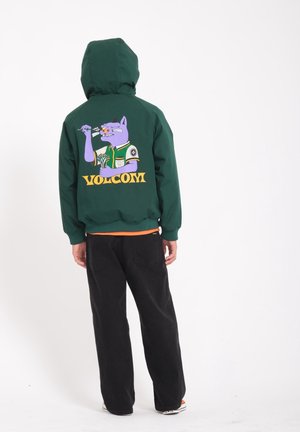 Groene capuchonjas met een afbeelding van een paarse cartoonkat op de achterkant, met de tekst "VOLCOM" en geribbelde boorden, gecombineerd met zwarte broek.