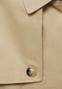 Beige trench coat fabric featuring a smooth texture, a large round button with a shiny finish, and a folded collar detail.