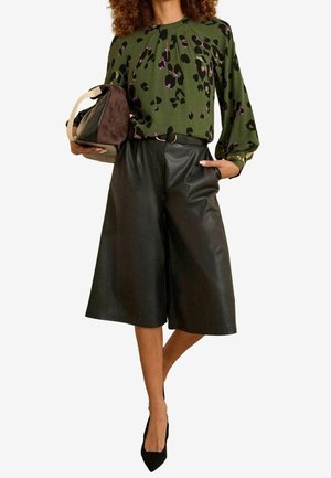 Vrouw die een olijfgroen blouse met luipaardprint draagt, zwarte leren culottes, zwarte hakken, en een bruine en beige handtas onder één arm houdt.