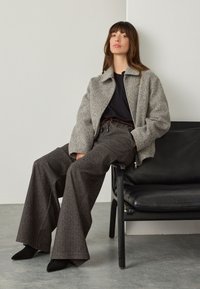 Gray textured jacket over a black top, paired with wide-leg dark patterned pants. Sitting on a black chair against a neutral background.