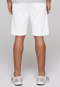 Pantalones cortos de algodón blanco con una silueta relajada, acabados con dobladillos enrollados. Combinados con zapatillas deportivas gris claro. Enfocándose en la vista trasera.