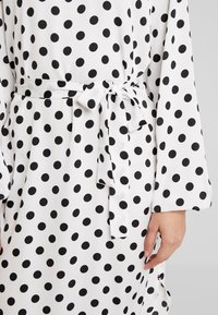 Weißes Kleid mit schwarzen Punkten; verfügt über einen gebundenen Taillengürtel, lange Ärmel und eine glatte Stofftextur. Auffälliges Muster im gesamten Kleid.