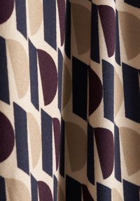 Tissu à motifs présentant des formes géométriques en beige, bleu marine, bordeaux et crème. La texture semble douce avec un léger éclat.