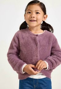 Häivähdyksellinen violetti neuletakki pyöreällä pääntiellä, nappikiinnityksellä edessä ja pitkillä hihoilla, kerrostettuna valkoisen, kukalliset yksityiskohdat sisältävän paidan päälle.