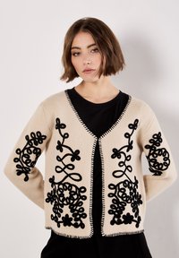 Beiger Cardigan mit schwarzem Blumenstickerei an den Ärmeln und vorne, mit V-Ausschnitt, offener Front und kontrastierendem Stich entlang der Kanten.