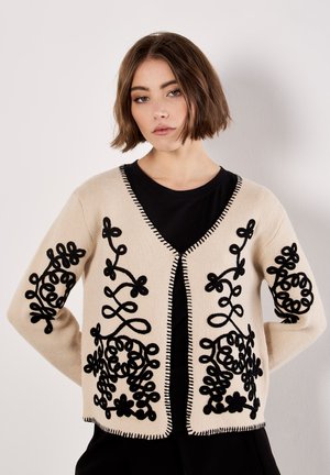 Beige cardigan with black floral embroidery on sleeves and front, featuring a V-neck, open front, and contrasting stitching along the edges.