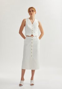 Gilet cropped in lino bianco con scollo a V e bottoni dorati, abbinato a una gonna bianca al ginocchio con placket frontale e sandali coordinati.